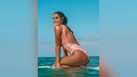 Smiling woman in a pink patterned one-piece swimsuit sitting on a surfboard in the ocean under a blue sky.