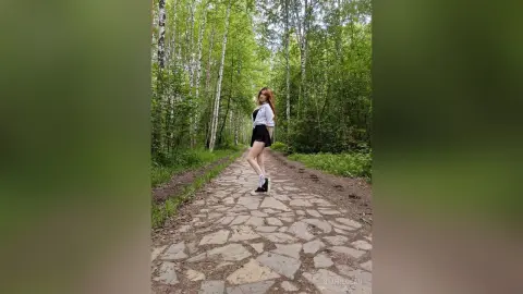 Redheaded woman in a white blouse and black shorts walking on a stone path through a lush green forest.