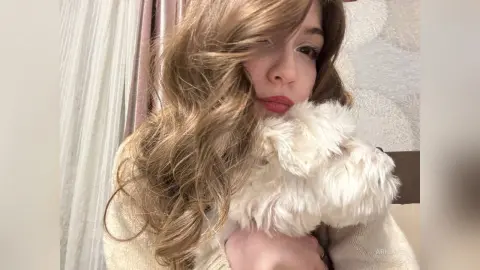 Blonde woman in a cozy cream sweater cuddling a fluffy white dog in a warm, indoor setting.