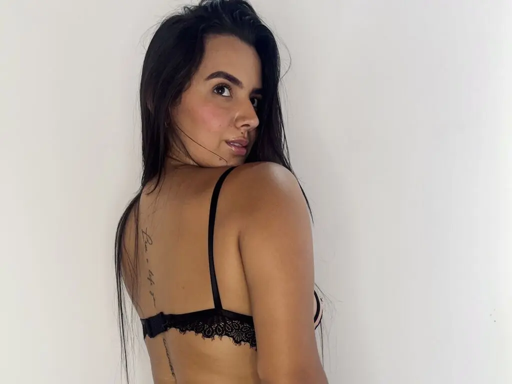 Young woman with long dark hair wearing a black lace bra looking over her shoulder against a white wall.