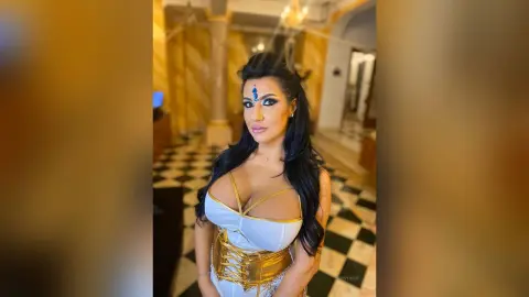 MissyJolie poses in a white and gold corset top, showing off her cleavage and long dark hair against a black and white tiled floor.
