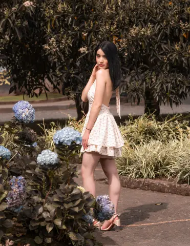 Young woman in a floral sundress standing by blue hydrangeas in a lush garden setting.