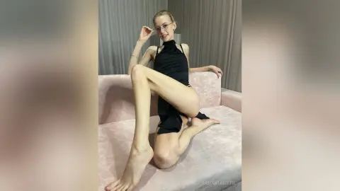 Blonde woman in a chic black halter dress posing playfully on a pink velvet sofa in a bright room.