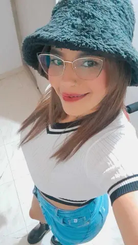 Smiling young woman in a white ribbed top, blue jeans, and a black fuzzy bucket hat wearing glasses.