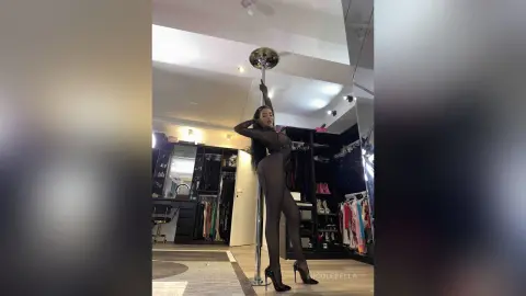 NicoleBella stands naked in a black bodysuit while holding onto a silver dance pole in a studio setting.