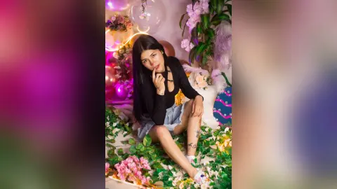 Young woman in a black long-sleeve top and denim shorts sitting amidst pink flowers and balloons in a dreamy setting.