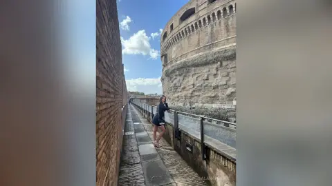 Stylish woman in a black dress standing on a narrow stone walkway beside a historic brick fortress under a blue sky.