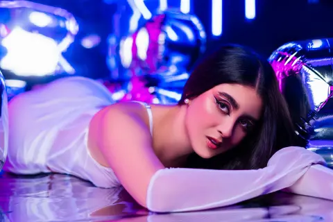 Glamorous woman in a white satin outfit posing amidst glowing pink and blue balloons in a dreamy studio setting.