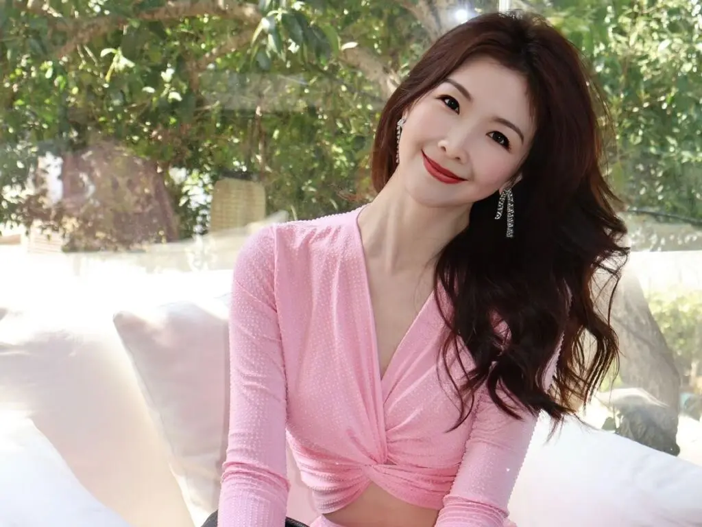 Smiling woman in a pink long-sleeve wrap top sitting on a white sofa in a bright outdoor garden setting.