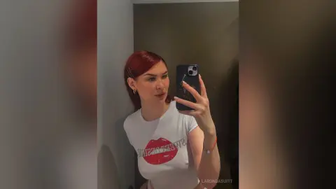 Redheaded woman in a white graphic t-shirt taking a casual mirror selfie in a dimly lit room.