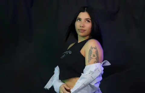 channeljonson — Young woman with long dark hair and arm tattoo wearing a black tank top against a dark studio background.