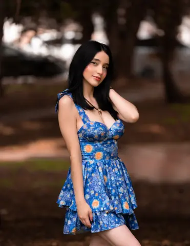 Young woman in a blue floral sundress posing in a dreamy, sunlit park setting.