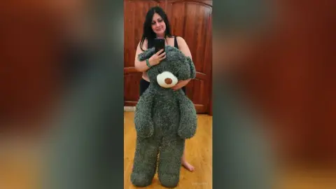 Smiling woman in a black tank top hugging a giant grey teddy bear in a cozy indoor setting.