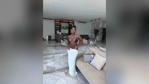 Confident woman in a black bikini and white maxi skirt posing in a modern, luxury marble living room.