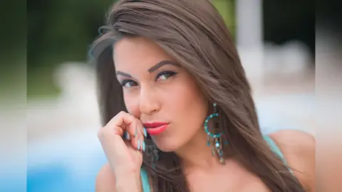 MontseFerrera poses with her hand near her mouth, wearing turquoise earrings and red lipstick against a soft blue background.