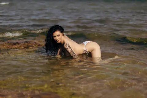 Tattooed woman in a white bikini relaxing in clear ocean water at a sunny beach.