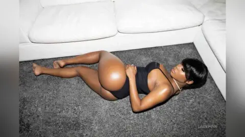 Curvy woman in a black bodysuit lying on a grey carpet in a bright, modern living room.