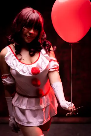 Smiling woman in a white ruffled clown costume holding a large red balloon in a moody, dark setting.