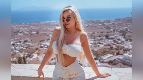 Claire poses outdoors in a white strapless crop top and matching bottoms, wearing sunglasses with a coastal town and sea in the background.