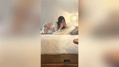 Tattooed woman in beige lingerie relaxing on a white bed in a cozy, dimly lit bedroom.
