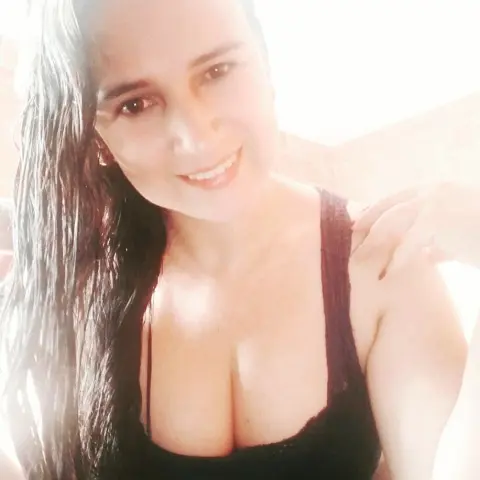 BrianaCharlot smiles while wearing a black tank top, showing her long dark hair and cleavage in a bright setting.