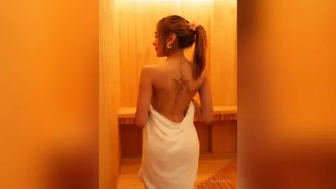 Woman in a white towel with back tattoos relaxing in a warm, wooden spa sauna setting.