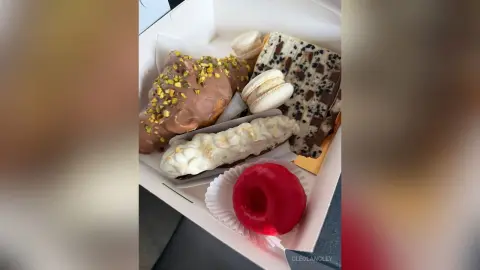 Assorted gourmet pastries including macarons, chocolate bark, and glazed donuts in a white gift box.