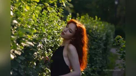 Redheaded woman in a black halter top posing amidst lush green garden foliage in soft sunlight.