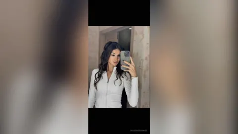 CatherineThomase — CatherineThomase takes a mirror selfie wearing a tight white long-sleeved shirt and long wavy dark hair.