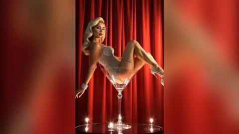Glamorous blonde woman in a sparkling silver bodysuit posing on a giant martini glass against red curtains.