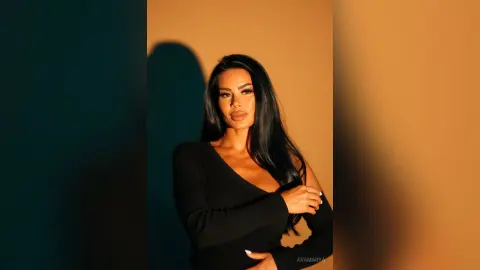 Elegant woman in a black long-sleeve top posing against a warm tan background with dramatic studio lighting.