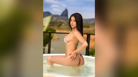 VickyPortman poses in a white bikini while sitting in a hot tub overlooking a tropical landscape.
