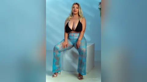 Blonde woman in a black bikini top and sheer blue floral trousers sitting on a white cube in a studio setting.