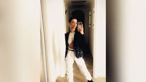 SofiaRoose poses in a mirror wearing a black leather jacket over a white top and white pants, holding her phone up to take a selfie.
