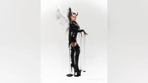 Woman in black latex bodysuit and horns with large white wings posing in a minimalist studio setting.