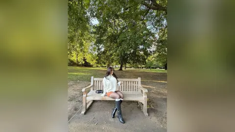 Young woman in a white sweater and black boots sitting on a park bench under lush green trees.
