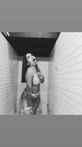 Tattooed woman in a sheer white outfit posing playfully in a white tiled shower room.