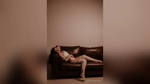 Elegant woman in white lingerie relaxing on a dark leather sofa in a minimalist, moody room.