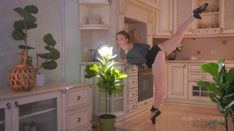 FlorenceClutter poses in a kitchen, leaning forward on a counter while lifting one leg high behind her, wearing a black striped top and black undies.