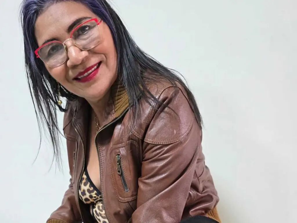 CelesteCorner smiles wearing red glasses, a brown leather jacket, and a leopard print top against a plain white background.