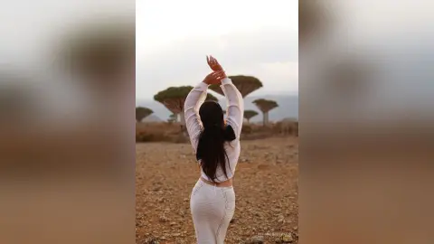 Woman in white outfit stretching her arms in a serene desert landscape with trees under a soft sky.