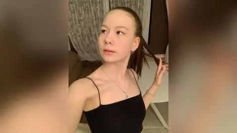 Young woman with slicked-back hair wearing a black camisole tank top in a cozy indoor setting.