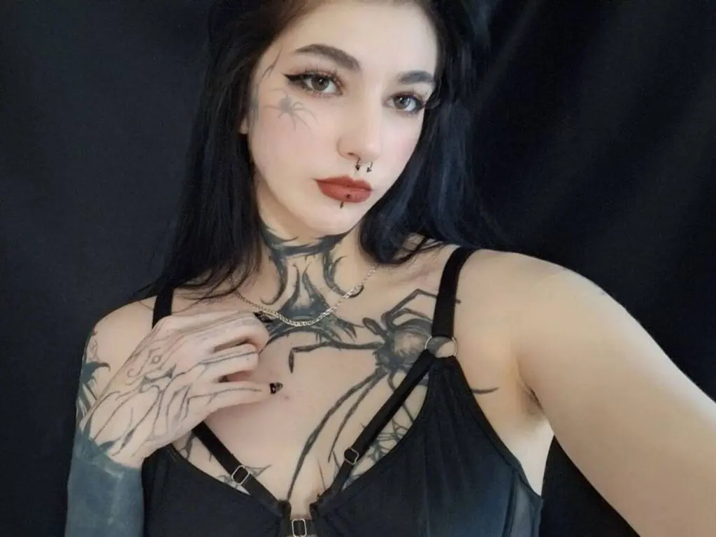 Woman with dark hair and facial tattoos wearing a black tank top against a dark background.
