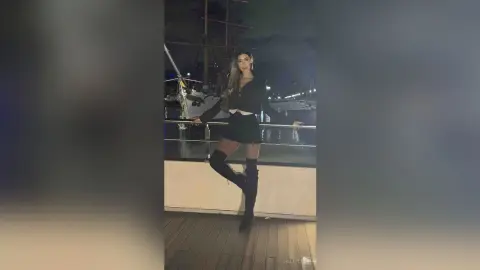 Stylish woman in a black crop top and mini skirt with over-the-knee boots posing on a moonlit boat deck.
