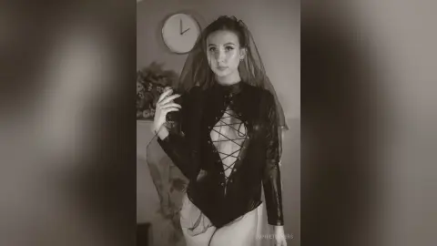 Moody black and white portrait of a woman in a dark lace corset and sheer veil indoors.