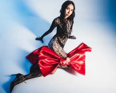 AilynHoney — AilynHoney poses in a black lace bodysuit and high heels against a blue background.