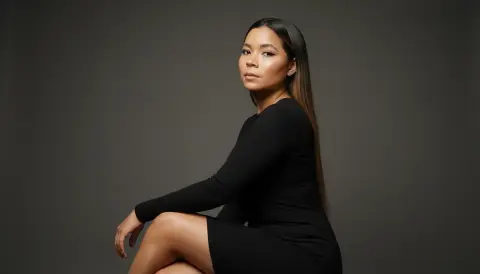 XinemaBonet poses in a long-sleeved black dress, looking over her shoulder with long straight hair against a dark grey background.