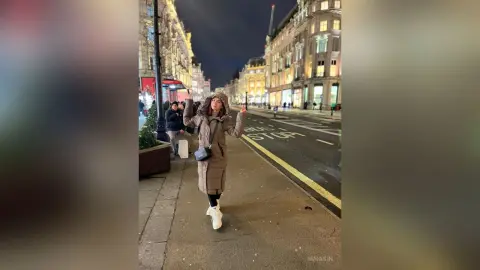 Young woman in a tan winter coat and white boots smiling on a city street at night.
