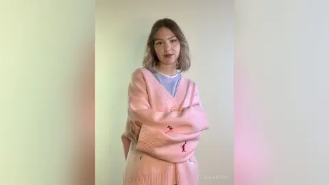 Young woman in a cozy pink oversized cardigan and light blue shirt posing against a neutral wall.