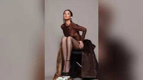 Elegant woman in a brown satin long-sleeve top and sheer tights posing glamorously on a chair against a grey backdrop.